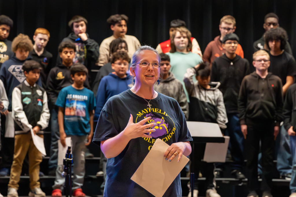 A teacher speaks to an audience, while the choir stands behind her on risers.