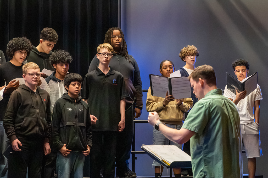 A section of middle school choir students sing while reading from music folders as a conductor leads from the front.