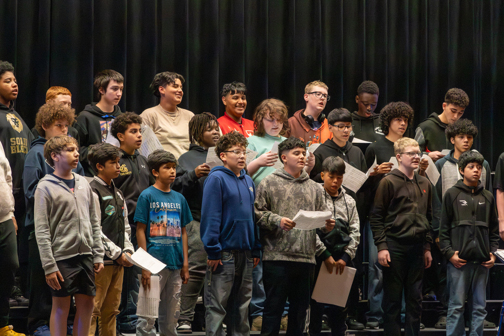 Middle school students stand on risers holding sheet music, singing together during a tenor-bass festival performance.