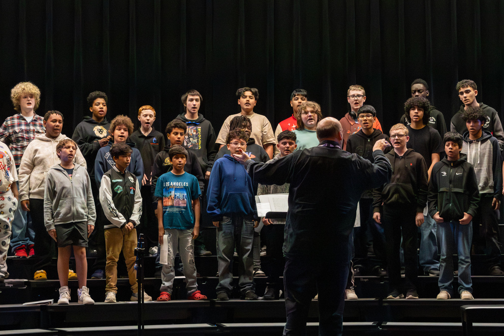Middle school students sing on stage while a conductor gestures with both hands, guiding the tenor-bass choir during a performance.