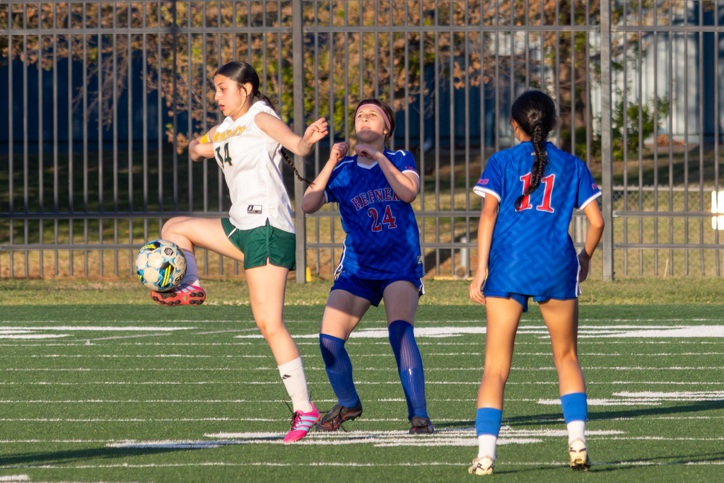 A Warriors player in white lifts her leg to control a bouncing ball while two Hefner players in blue defend nearby.