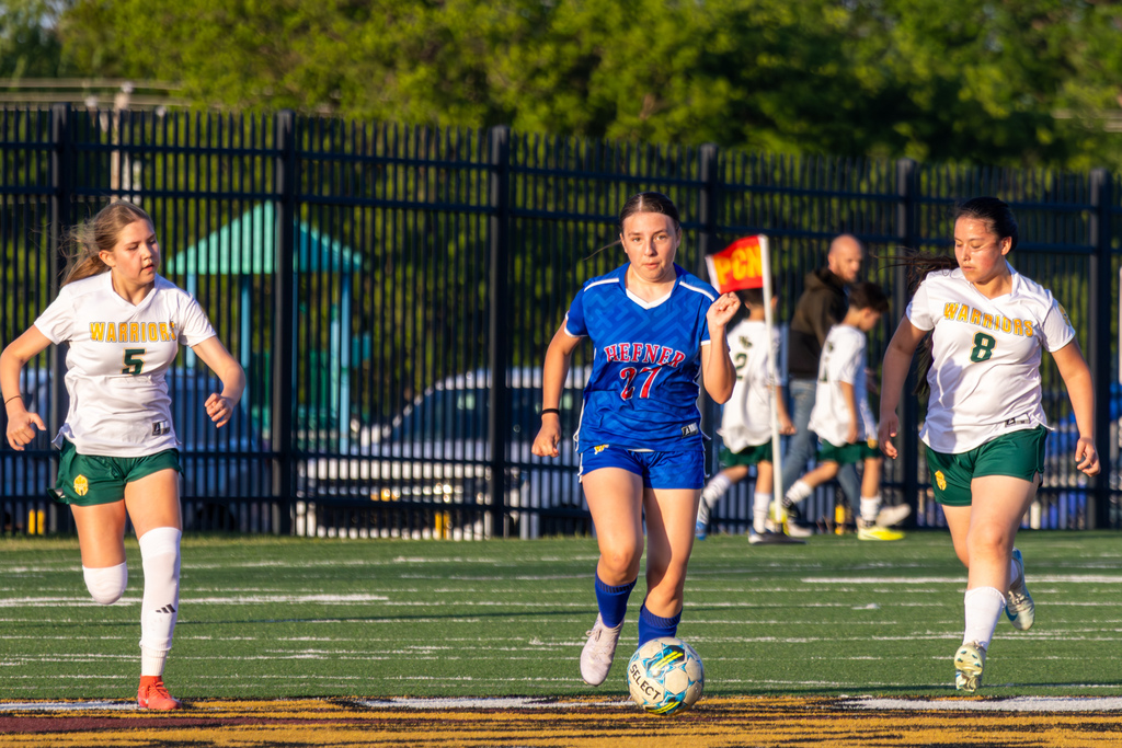 A Hefner player in blue dribbles the ball up the center of the field while two Warriors players in white close in from either side.