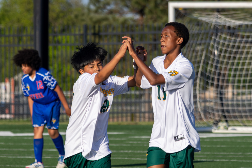 Two Warriors teammates in white jerseys meet for a quick high-five near the goal while a Hefner player in blue walks in the background.