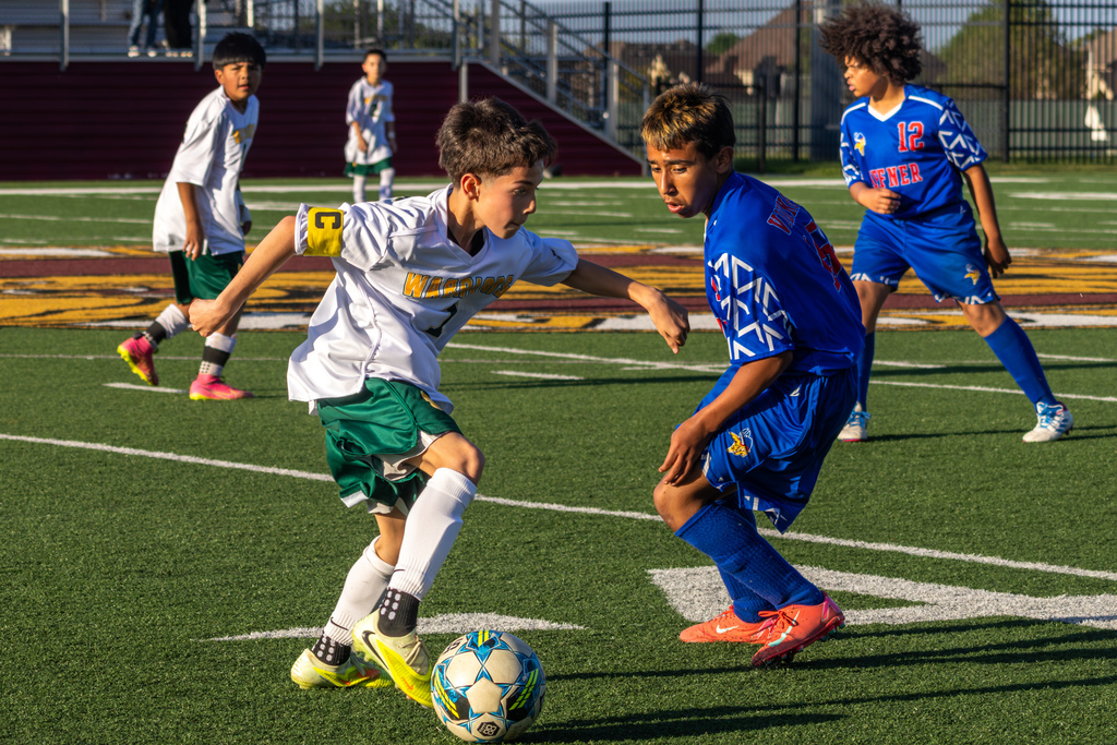 Two middle school boys compete for the ball at midfield, one in a white Warriors jersey dribbling past a defender in a blue Hefner uniform as teammates watch behind them.