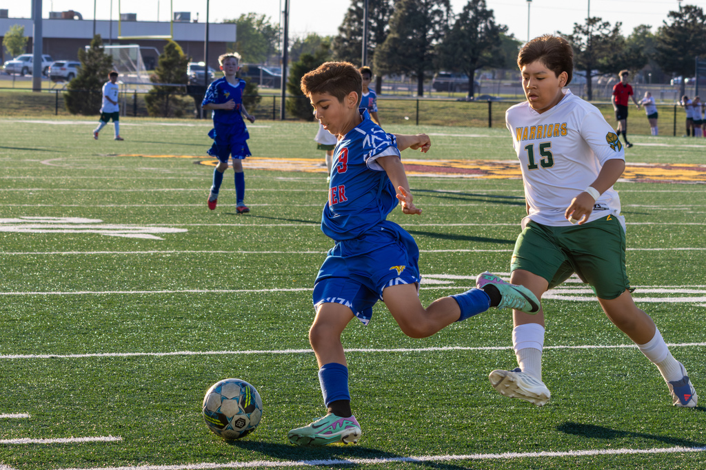 A Hefner player in blue dribbles downfield while a Warriors defender in white chases closely, both focused on the ball during live play.