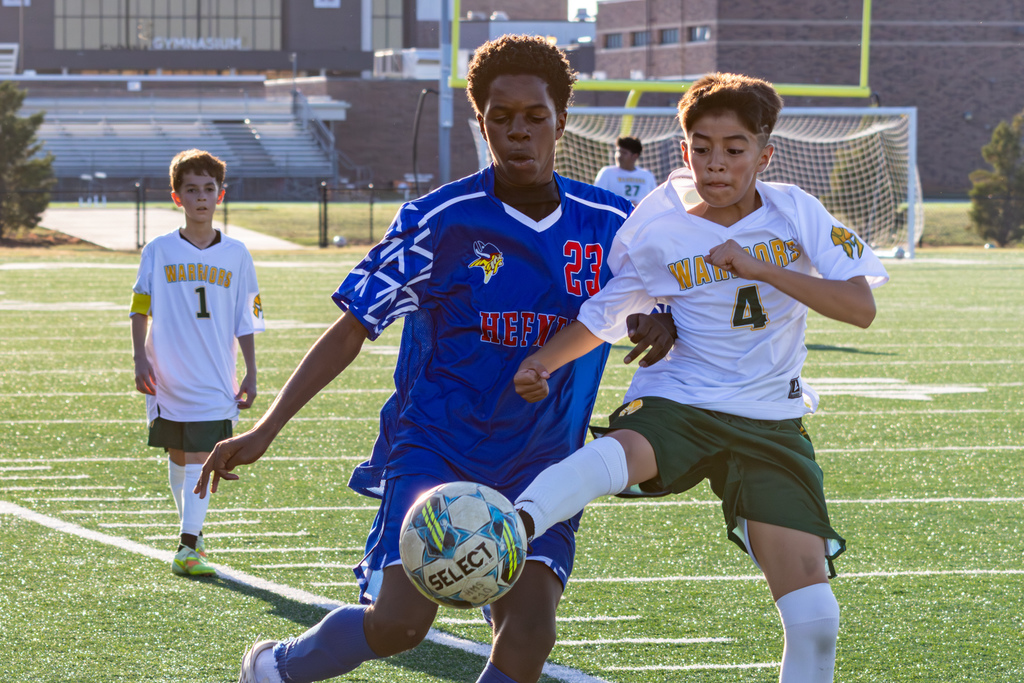 Two players sprint side by side toward the ball, one in a blue Hefner jersey and one in a white Warriors jersey, both reaching with their feet to gain control.