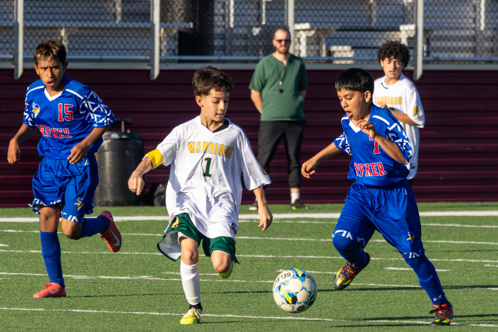 A Warriors player in white dribbles between two Hefner defenders in blue, all three focused on the ball during an active play.