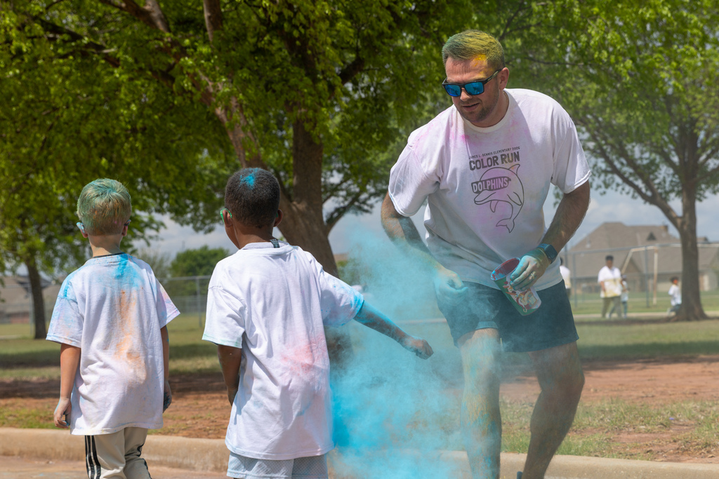 An adult tosses blue powder toward two young students walking along the course, creating a cloud of color around them.