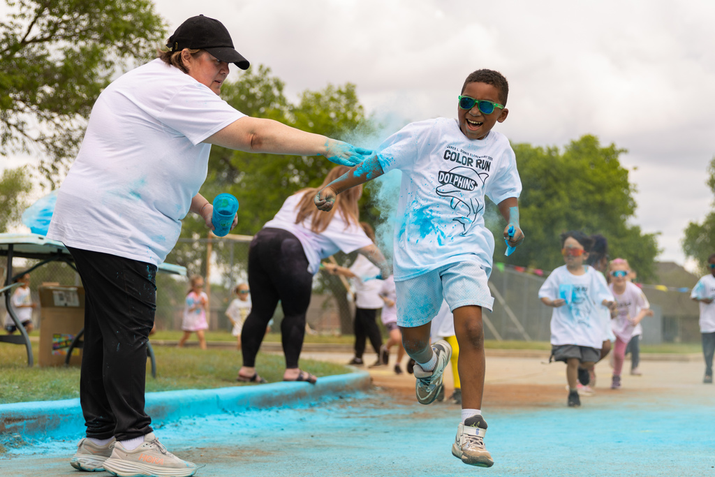 A student runs past as an adult throws blue powder onto his shirt, with other students continuing along the Color Run route in the background.