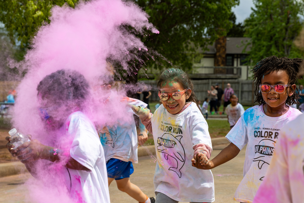A burst of pink powder fills the air as a group of students runs through, one student smiling while holding hands with a classmate.
