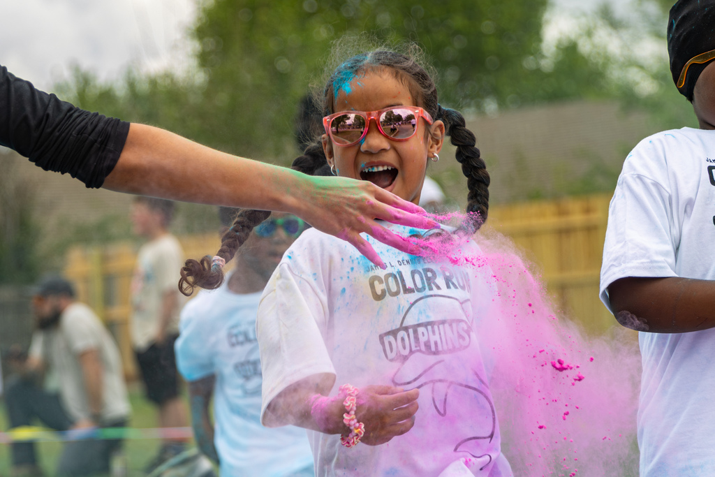 A student in pink sunglasses smiles with her mouth open as an adult tosses bright pink powder across her shirt during the Color Run.
