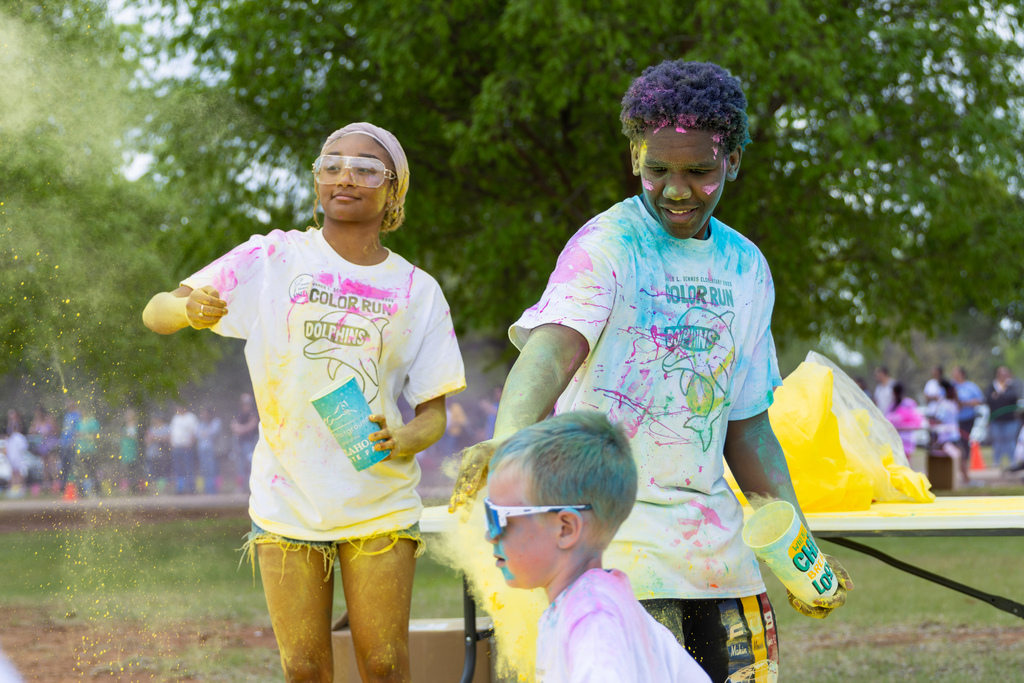 Two older students at a color station toss yellow powder onto a younger student passing by, all covered in colorful dust.