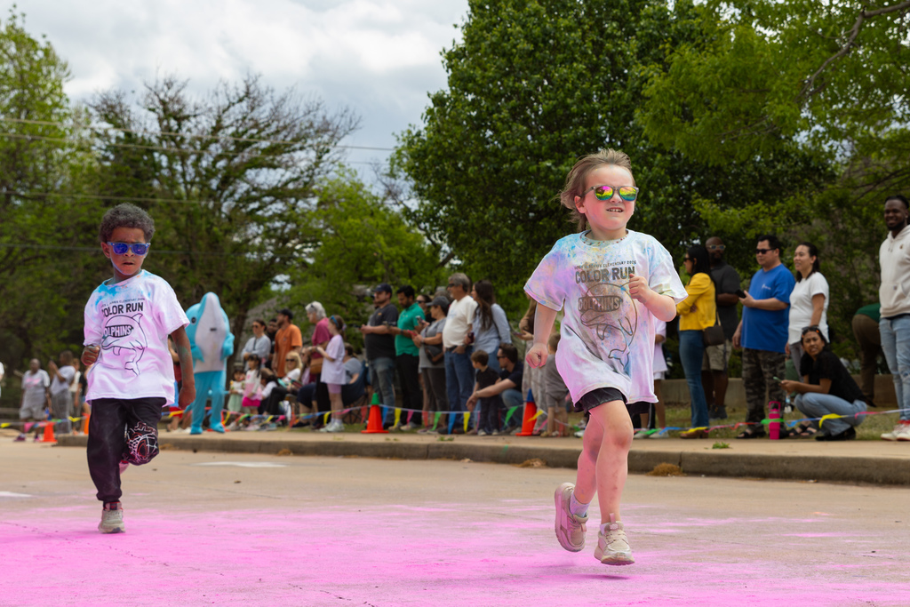 Two young students run side by side on a powder-covered path, with families and spectators lined up along the sidewalk watching.