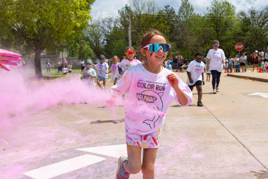 A student wearing reflective sunglasses runs toward the camera as pink powder is tossed across the path and other runners follow behind.