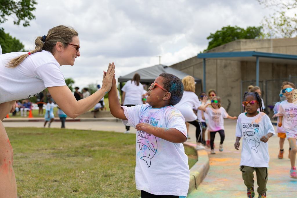A teacher and a student share a high-five along the course while other students in white Color Run shirts jog behind them.