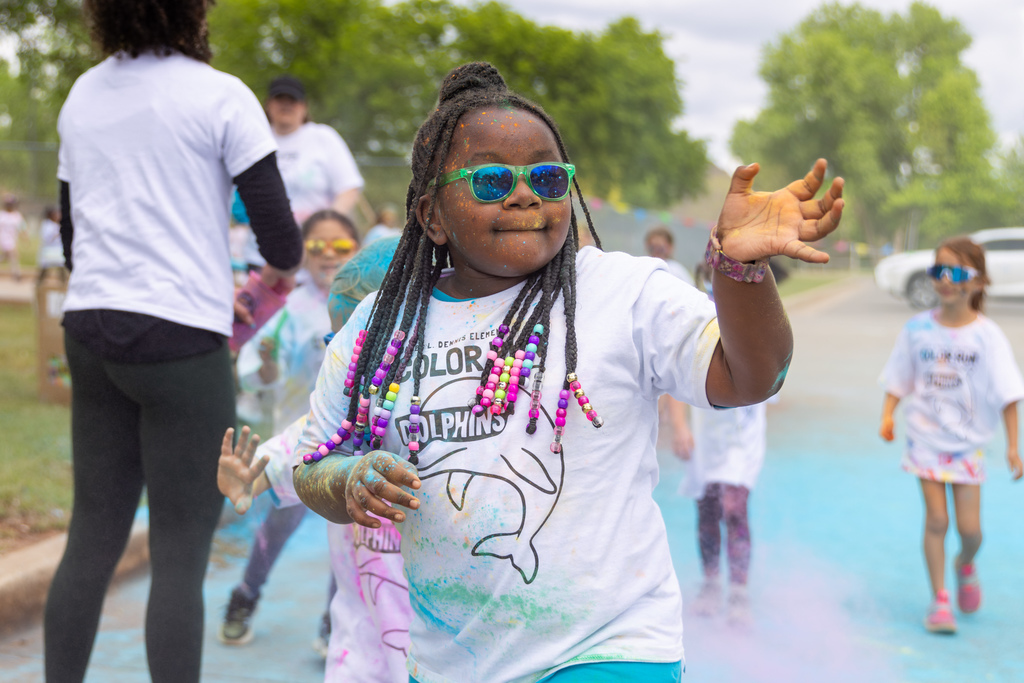 An elementary student wearing sunglasses and beaded braids runs through a cloud of colored powder, waving her hand as other students follow behind on the course.