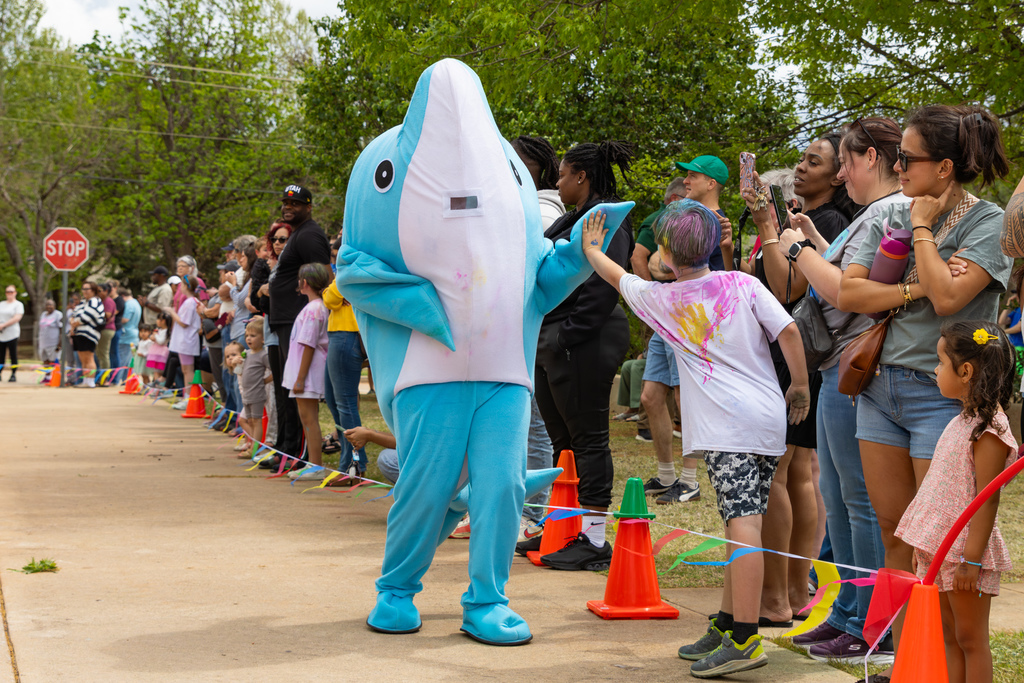A student reaches out to high-five a person in a blue dolphin mascot costume while a crowd of families stands along the course.