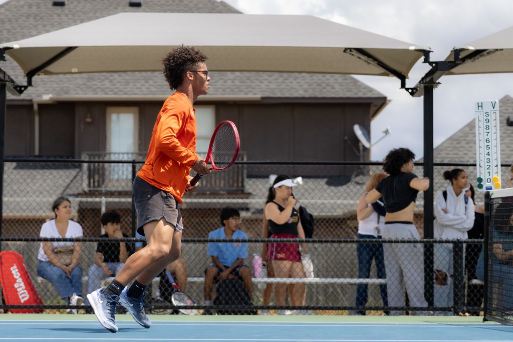A Putnam City High tennis player bounces lightly on his feet in a ready stance while audience members watch.