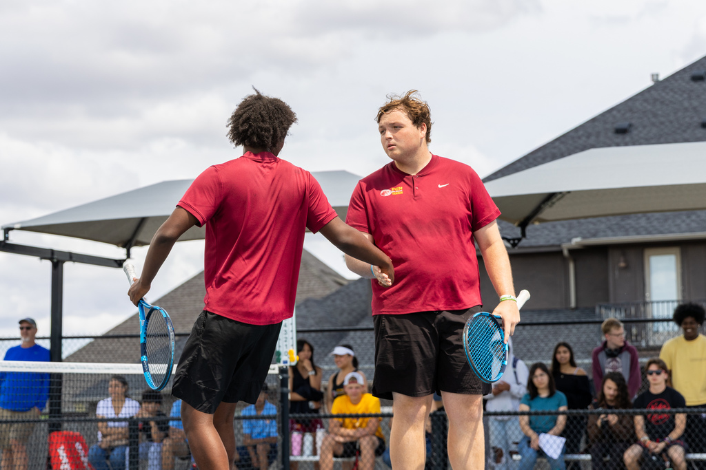Two Putnam City North tennis players meet at the net for a handshake during a match while spectators watch in the background.