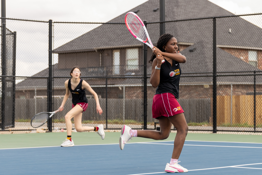 A Putnam City North girls tennis player follows through on a backhand shot while her doubles partner tracks the ball behind her on an outdoor court.