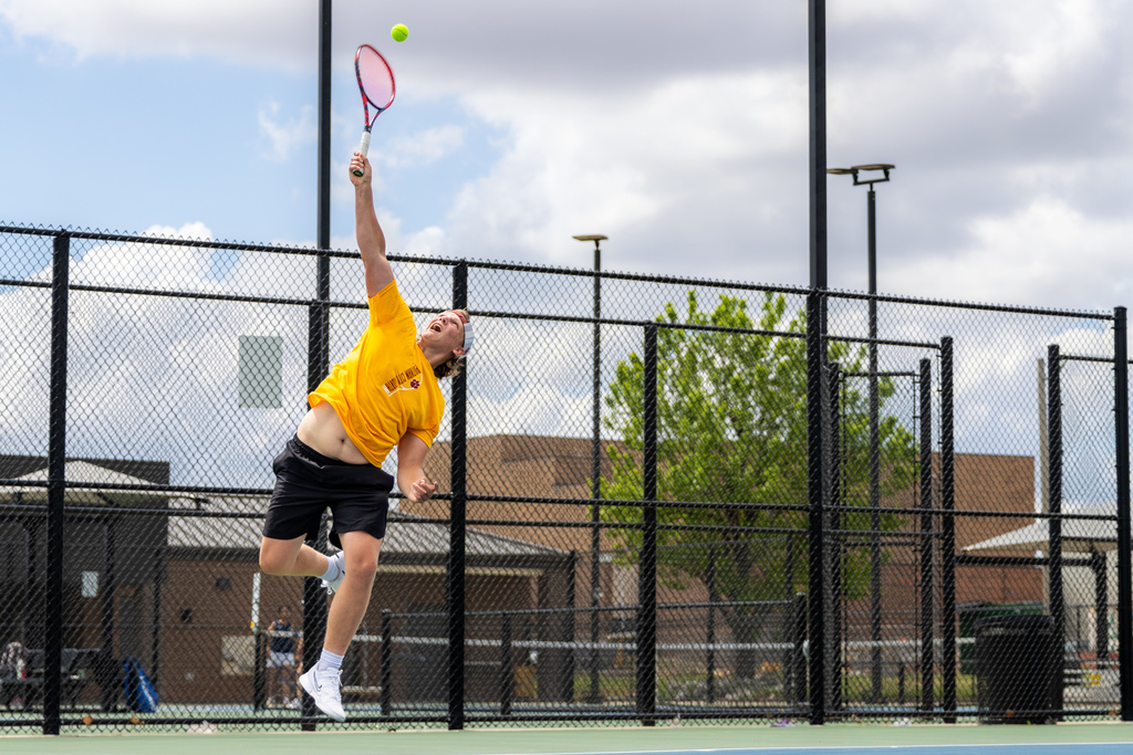 A Putnam City North tennis player jumps into an overhead serve, arm fully extended toward the ball against a bright sky.