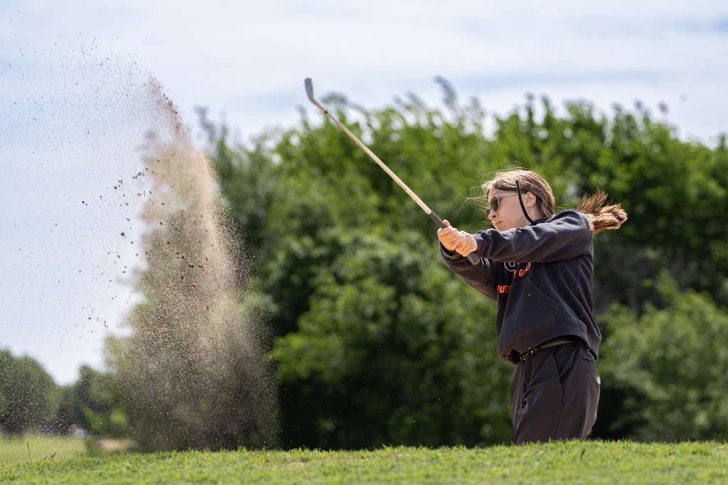 Student golfer hits out of a sand bunker, with sand spraying into the air during a tournament round.
