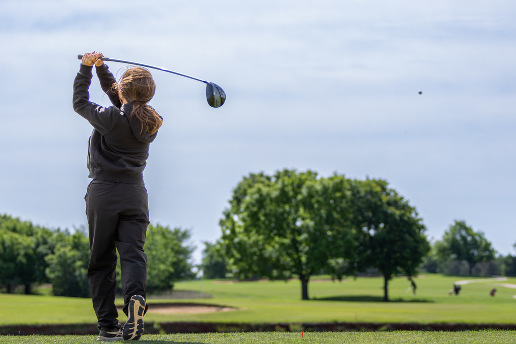 Student golfer follows through on a drive, watching the ball in flight over the fairway.