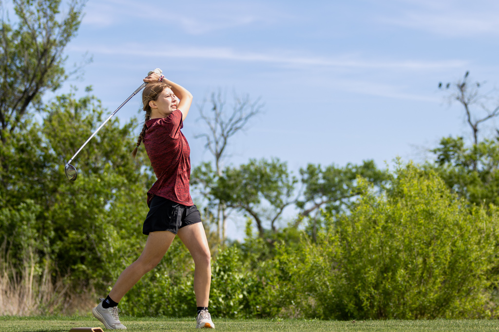 Student golfer in a red shirt completes a full swing on the tee box during regional play.