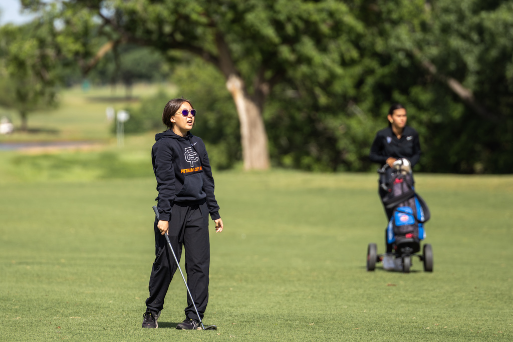 Student golfer walks the fairway holding a club, with another golfer and push cart in the background.