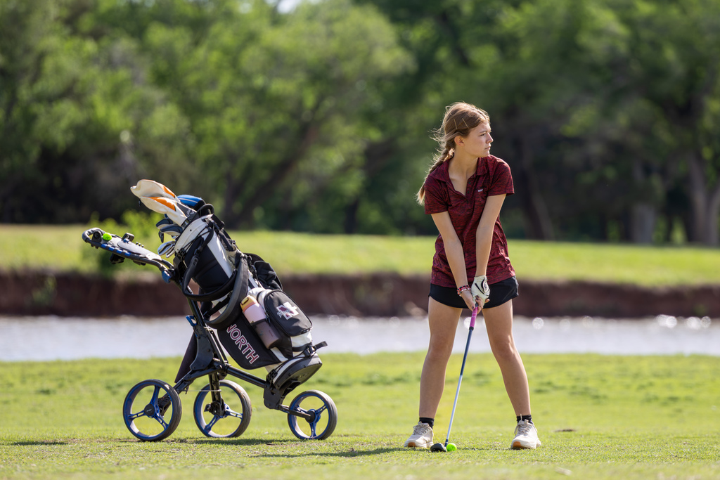 Student golfer stands at address beside a golf bag and push cart, preparing for a shot.