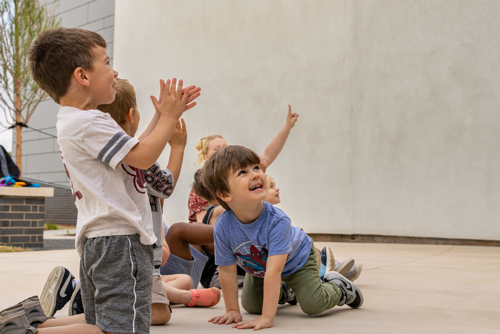 Young children sit and kneel on a concrete courtyard, clapping and looking upward with excitement as something catches their attention just out of frame.