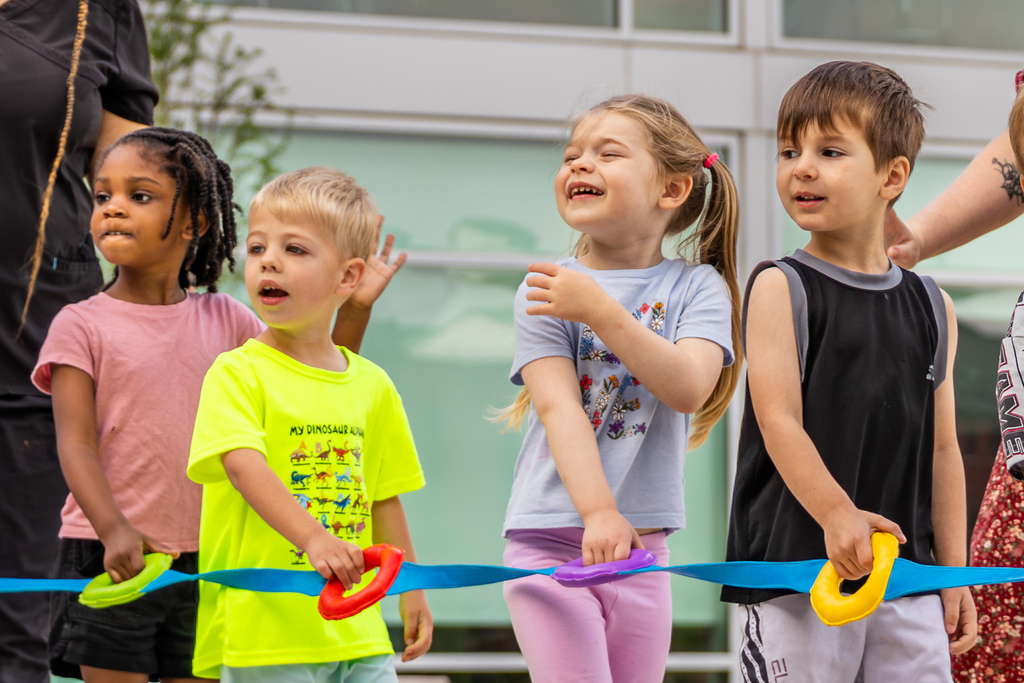 Several children stand side by side holding colorful rings on a blue ribbon, looking off to the side as if waiting for instructions or watching an activity.