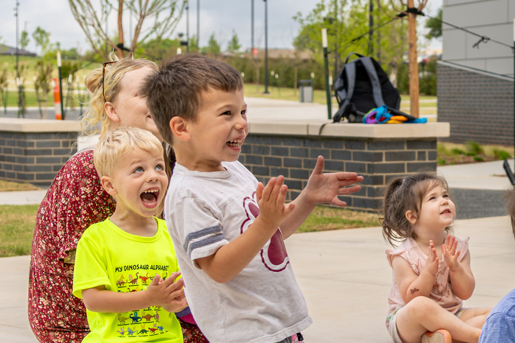 Children smile, clap, and react with excitement, gathered together outdoors near a brick planter and greenery.