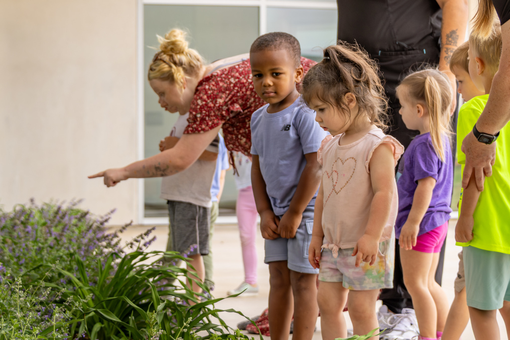Children stand in a row near a garden bed while an adult points toward plants, guiding them as they observe something in the greenery.