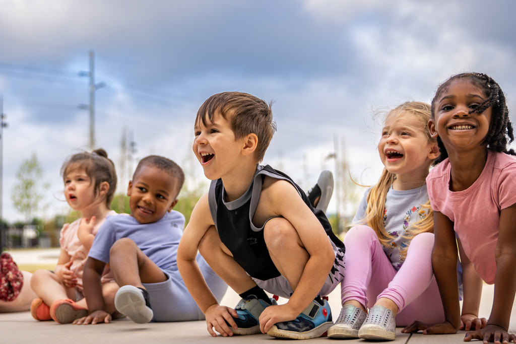 A group of young children sit on the ground in a line, smiling and laughing as they look ahead, with cloudy skies and trees in the background.