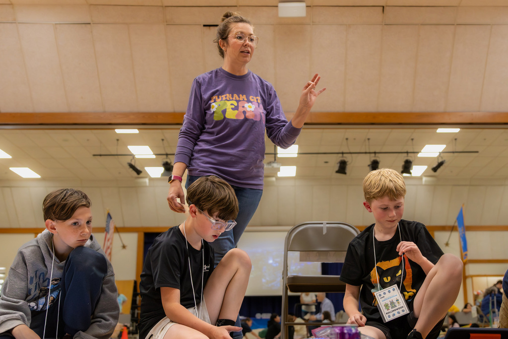 A teacher stands over a group of seated students, giving instructions with her hand raised as the students prepare their robotics materials on the floor.