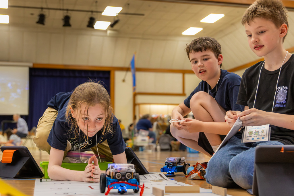Three students lie and kneel on a gym floor, focused on a small robot moving across a printed mat while holding controllers and pieces nearby.