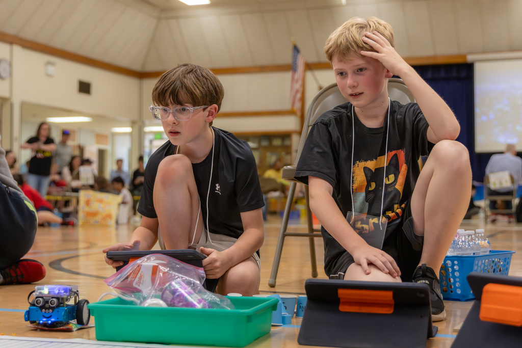 Two students in matching shirts lean in toward a robot and small cups on a mat, watching closely as it moves through the course.