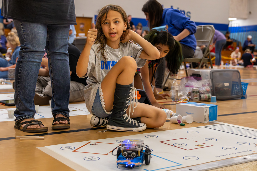 A student sits cross-legged and gives a thumbs up beside a robot on a competition mat, with supplies and teammates nearby.