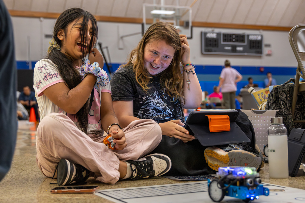 Two students sit on the floor laughing and reacting to a robot’s movement while using a tablet controller.