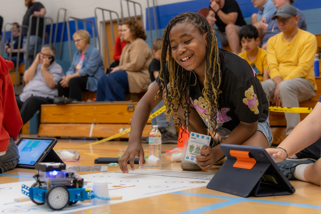 A student smiles while reaching toward a robot on the floor, with spectators seated on bleachers behind her watching the competition.