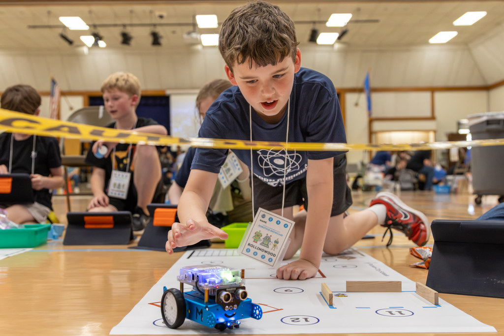 A student reaches toward a moving robot on a mat while concentrating, with other teams working in the background.