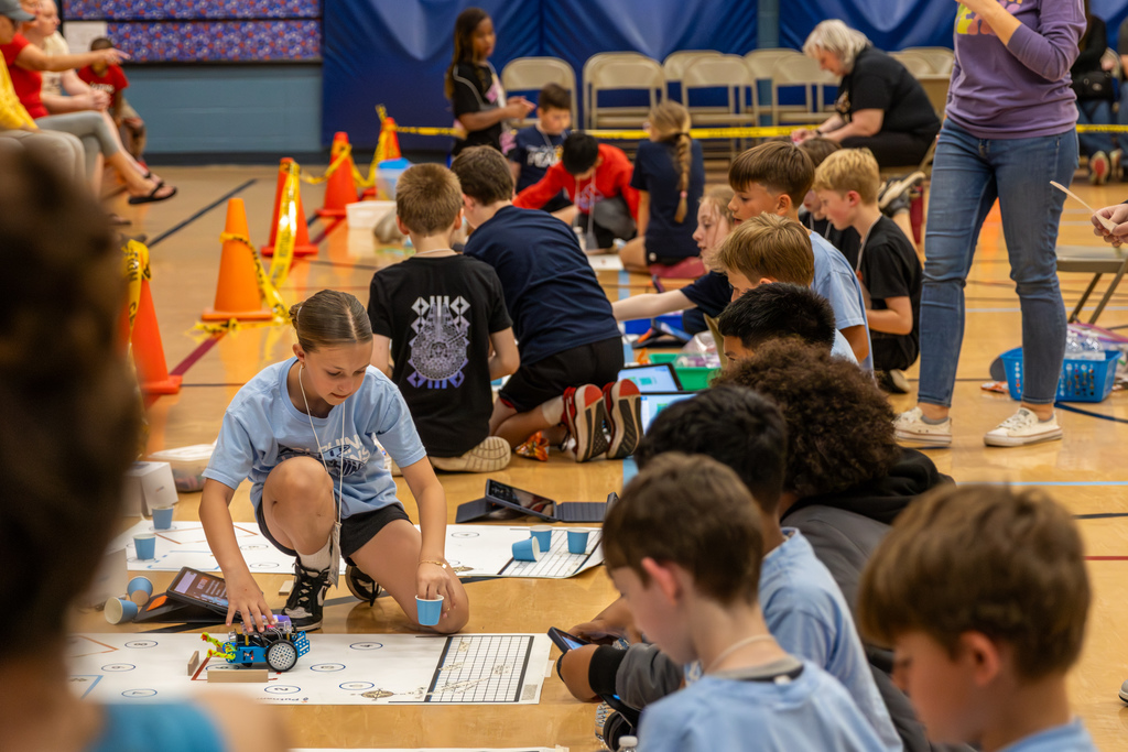 A wide view of multiple student teams spread across a gym floor, each working with robots and tablets at their own competition stations.