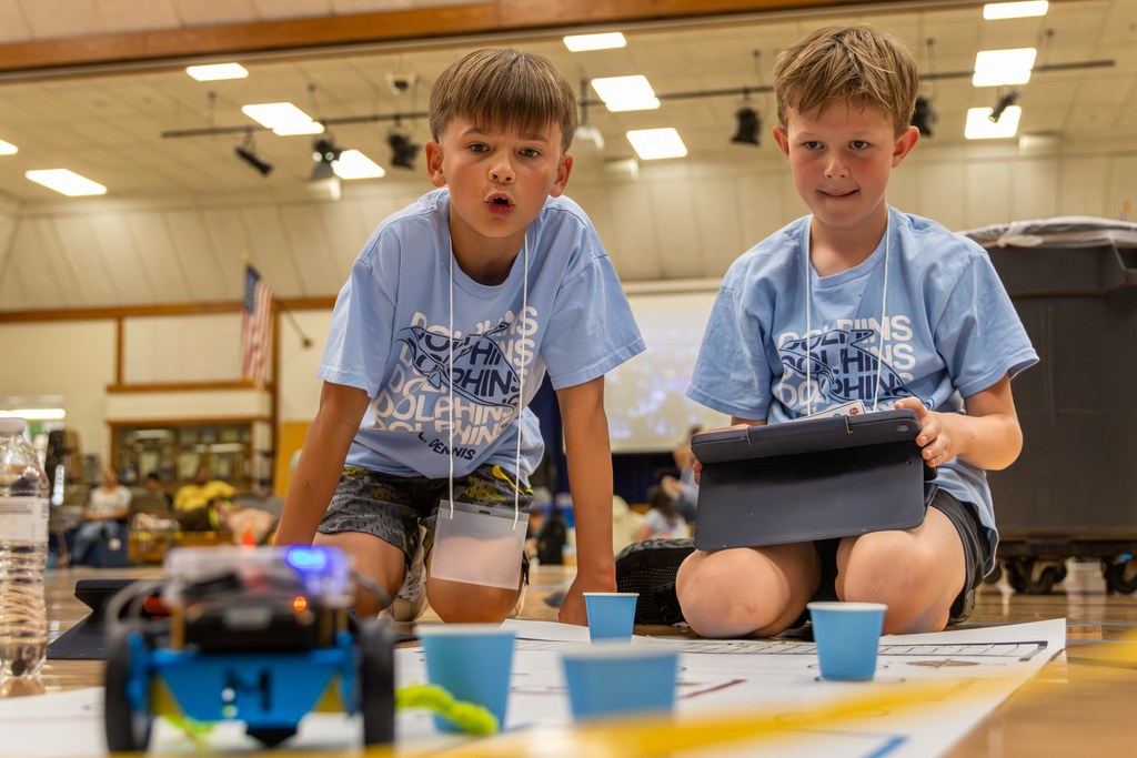 Two students kneel close to a robot and tablet, concentrating as they adjust their program before a run.