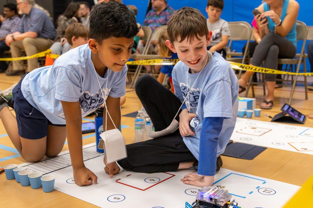 A student reaches toward a moving robot on a mat while concentrating, with other teams working in the background.