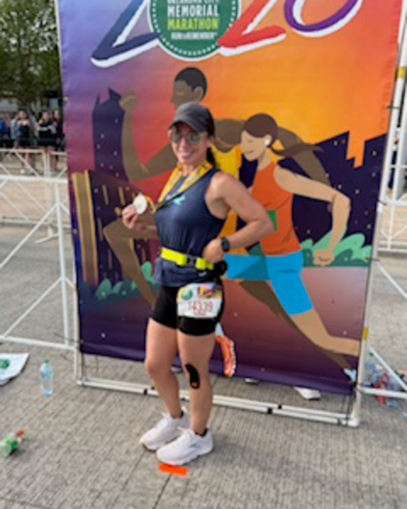 A runner poses in front of a colorful marathon sign, holding her medal and standing in running gear near the finish area.