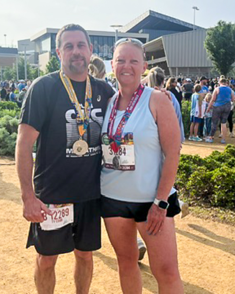 Two runners stand together with medals around their necks, smiling after finishing the race near the event grounds.