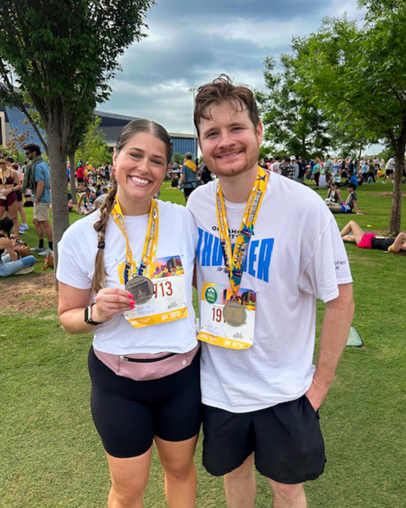 Two runners stand side by side on a grassy area, smiling and showing their medals with a large crowd gathered behind them.