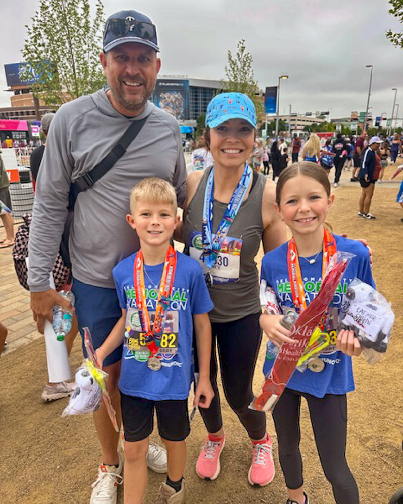 A family of four poses together after the race, all smiling and wearing medals, with event crowds and buildings in the background.