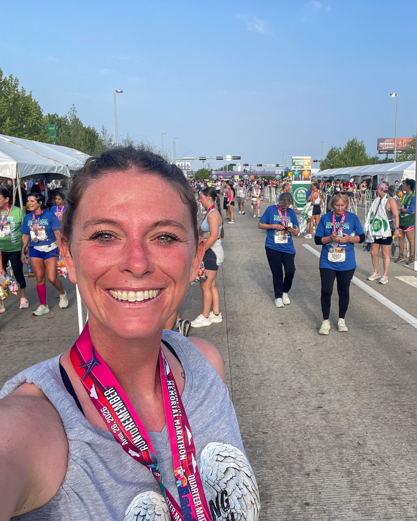 A runner takes a selfie at the finish area, smiling with a medal around her neck as other participants and tents line the road behind her.